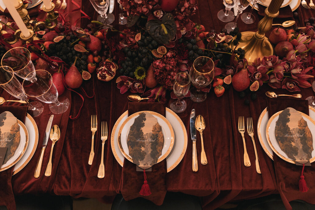 Baroque Wedding Feast setting gold and white plates and cutlery on a maroon tablecloth with fruit and charcuterie